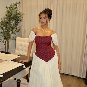 Puede incluir: Un vestido formal con un corpiño estampado en rojo y negro, mangas caídas y una falda color crema. El vestido tiene cintura alta y falda en capas. La mujer lleva un collar y pendientes.