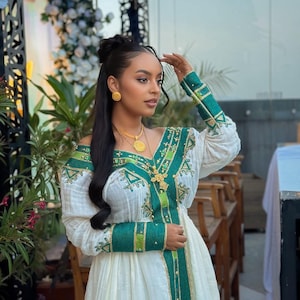 Puede incluir: Un vestido blanco con bordados verdes y dorados. El vestido tiene cuello en V y mangas largas con detalles verdes y dorados. La mujer lleva pendientes de oro y un collar de oro.