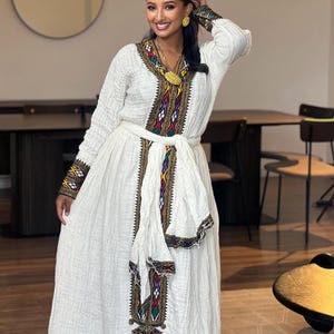 Puede incluir: Un vestido blanco con mangas largas y bordados intrincados en una variedad de colores, incluyendo rojo, verde y dorado. El vestido presenta un cinturón decorativo y se usa con joyas de oro. Parece ser una prenda tradicional.