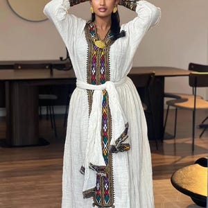 Puede incluir: Un vestido blanco con bordados coloridos y un cinturón a juego. El vestido presenta intrincados patrones en rojo, verde, amarillo y negro. Las mangas y el escote también están adornados con diseños detallados. Es una prenda tradicional.