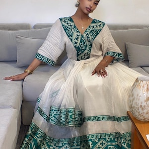 Puede incluir: Un vestido color crema con detalles bordados en verde. El vestido presenta un escote en V y mangas anchas. El bordado verde se concentra alrededor del escote, las mangas y el dobladillo. El vestido está ceñido a la cintura.