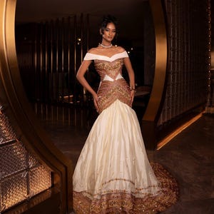 Puede incluir: Un vestido formal con hombros descubiertos, con una falda color crema y un corpiño con bordados dorados y marrones. El vestido presenta una silueta de sirena y detalles intrincados.