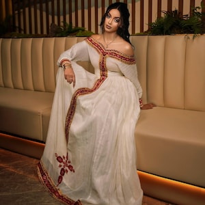 Puede incluir: Un vestido blanco con hombros descubiertos y ribetes rojos y dorados. El vestido tiene mangas largas y una falda fluida. El ribete presenta patrones intrincados. El vestido lo lleva una persona sentada en un sofá beige.