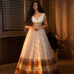 Puede incluir: Un vestido de fiesta blanco con escote en V y falda fluida. El vestido presenta detalles bordados dorados y marrones alrededor de la cintura y el dobladillo. La modelo lleva pulseras y un collar dorados.