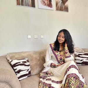 Puede incluir: Una mujer con un vestido blanco con intrincados bordados en tonos morados, dorados y negros. El vestido tiene mangas largas y un patrón detallado en los bordes. Está sentada en un sofá beige con cojines decorativos, sonriendo.