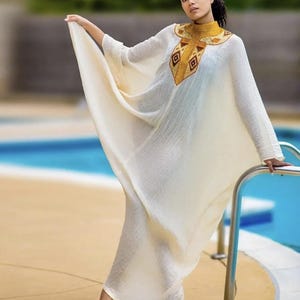 May include: A cream-colored caftan dress with long sleeves and a decorative gold and brown embroidered neckline. The dress is flowing and elegant, paired with gold heels. The background includes a pool and a sunny outdoor setting.