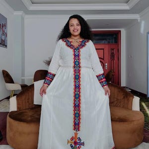 May include: A white dress with long sleeves and colorful embroidered details. The dress features a detailed pattern down the front and on the sleeves, with a cross-like design at the bottom. The dress is worn by a person.