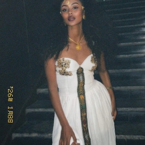 Puede incluir: Una mujer con un vestido blanco con bordados dorados y una abertura alta. El vestido presenta un panel vertical decorativo y está adornado con joyas doradas, incluyendo un collar, pendientes y una diadema. En el fondo, una escalera.