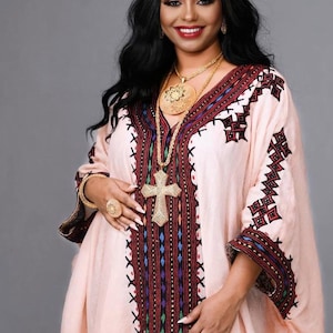 Puede incluir: Una mujer vistiendo un vestido tradicional etíope de color rosa claro con bordados intrincados en rojo, negro y azul. Lleva un collar de oro con un gran colgante y un colgante de cruz de oro.