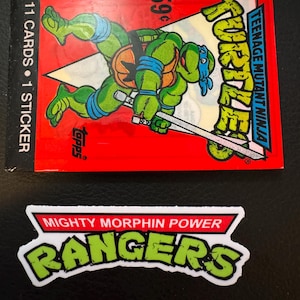 Può includere: Un pacchetto rosso di figurine dei Teenage Mutant Ninja Turtles con un'immagine a cartoni animati di una tartaruga. Il pacchetto include 11 figurine e 1 adesivo. È presente anche un adesivo dei Mighty Morphin Power Rangers.