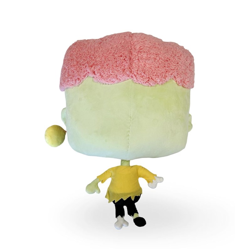 May include: A plush toy shaped like a green zombie with pink hair. The zombie is wearing a yellow shirt and black pants. The toy is sitting on a white background.