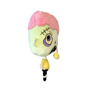 May include: A plush toy shaped like a zombie with pink hair, one eye, and a stitched mouth. The toy is wearing a yellow shirt and black pants.