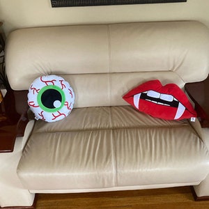 May include: Two decorative throw pillows on a cream colored couch. One pillow is shaped like an eye with a green iris and red veins. The other pillow is shaped like a red mouth with white teeth.