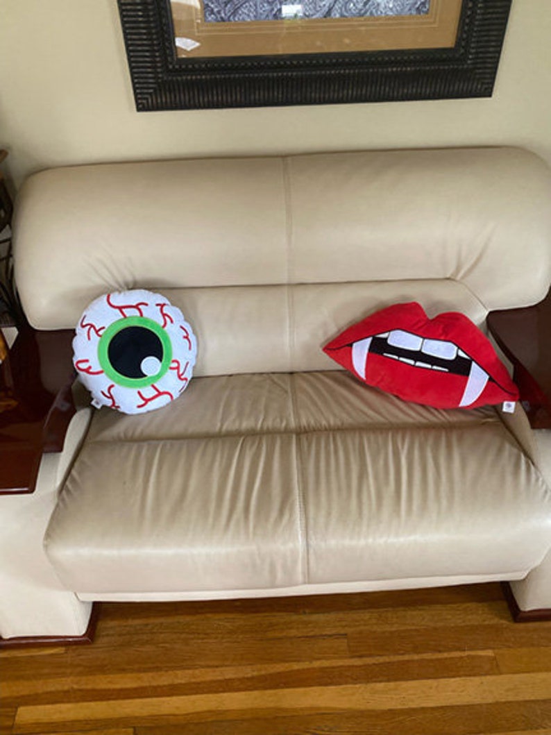 May include: Two decorative throw pillows on a cream colored couch. One pillow is shaped like an eye with a green iris and red veins. The other pillow is shaped like a red mouth with white teeth.