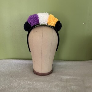 Könnte beinhalten: Ein schwarzer Haarreif, der mit einer Reihe flauschiger Pompons in einer bestimmten Farbabfolge verziert ist. Die Pompons sind von links nach rechts in Schwarz, Lila, Weiß, Gelb und Schwarz angeordnet und bilden ein strukturiertes Haaraccessoire.