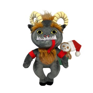 May include: A gray plush toy with horns, a red scarf, and a long tongue. The toy is holding a gingerbread man with a skull face and a red Santa hat.