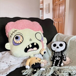 May include: Two plush toys, a green zombie-like character with a pink mohawk and a black skeleton with a skull head, are sitting on a couch with a white crocheted blanket.