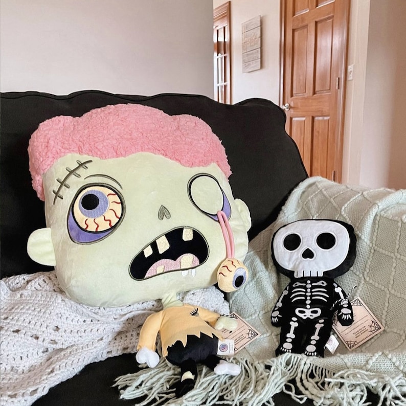 May include: Two plush toys, a green zombie-like character with a pink mohawk and a black skeleton with a skull head, are sitting on a couch with a white crocheted blanket.