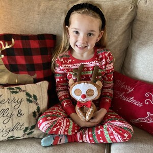 Op de afbeelding: Een jong meisje in rood-witte kerstpyjama zit op een bank en houdt een pluche rendier speelgoed vast. Het rendier heeft een skeletontwerp en een strikje. De bank heeft kussens met kerstthema's.