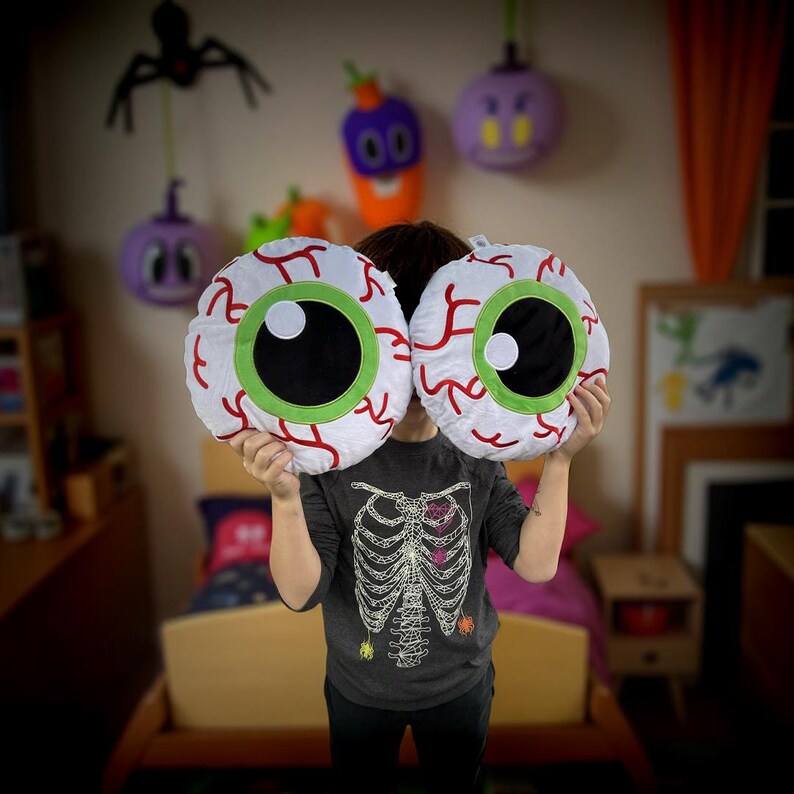 May include: Two plush white eye pillows with red veins and green irises. The pillows are being held in front of a child's face.