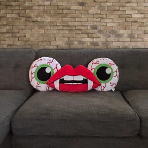 May include: Three decorative pillows on a gray couch. The pillows are shaped like eyes and a mouth with fangs. The eyes are white with green irises and black pupils. The mouth is red with white teeth.