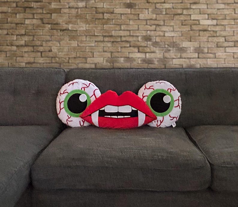 May include: Three decorative pillows on a gray couch. The pillows are shaped like eyes and a mouth with fangs. The eyes are white with green irises and black pupils. The mouth is red with white teeth.