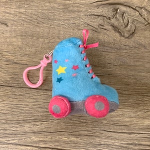 Op de afbeelding: Een blauwe pluche rolstoelschlüsselhanger met roze wielen en een roze en witte touwtje. De rolstoel heeft gele, groene en roze sterren.