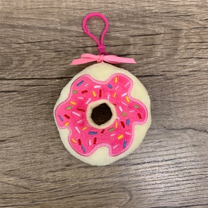 May include: A pink and white donut-shaped plush keyring with colourful sprinkles. The keyring has a pink ribbon and a pink clip.
