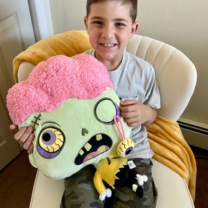May include: A child holding a green and pink plush zombie toy with a yellow and black plush zombie character attached. The zombie toy has a pink brain, one eye, and a stitched mouth.