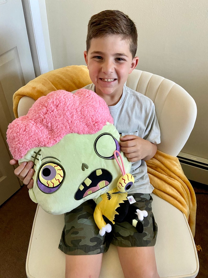 May include: A child holding a green and pink plush zombie toy with a yellow and black plush zombie character attached. The zombie toy has a pink brain, one eye, and a stitched mouth.