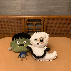 Op de afbeelding: Drie pluche speelgoeddieren op een beige beddengoed. Een groen monster met een brede grijns, een witte geest met &eacute;&eacute;n groot oog en een klein grijs monster met drie poten.