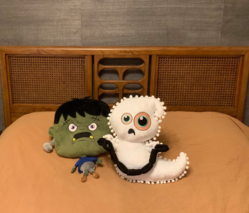Op de afbeelding: Drie pluche speelgoeddieren op een beige beddengoed. Een groen monster met een brede grijns, een witte geest met &eacute;&eacute;n groot oog en een klein grijs monster met drie poten.