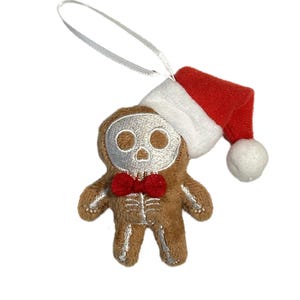 May include: A brown plush gingerbread man ornament with a skeleton face and rib cage design, wearing a red bow tie and a red and white Santa hat. The ornament has a silver string for hanging.