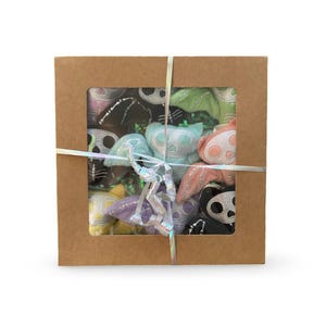 May include: A square brown cardboard gift box with a central rectangular window containing multiple small, plush bat-shaped toys in various pastel colours including pink, blue, green, yellow, and purple, tied with a shiny iridescent ribbon.