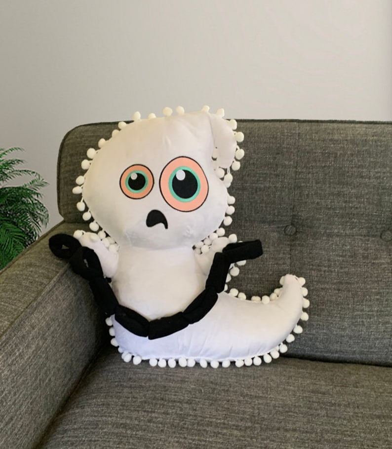 Op de afbeelding: Wit pluche kussen in de vorm van een spook met zwarte ogen en een fronsende mond. Het spook heeft een zwarte ketting om zijn nek en witte pompoentjes rond de randen.