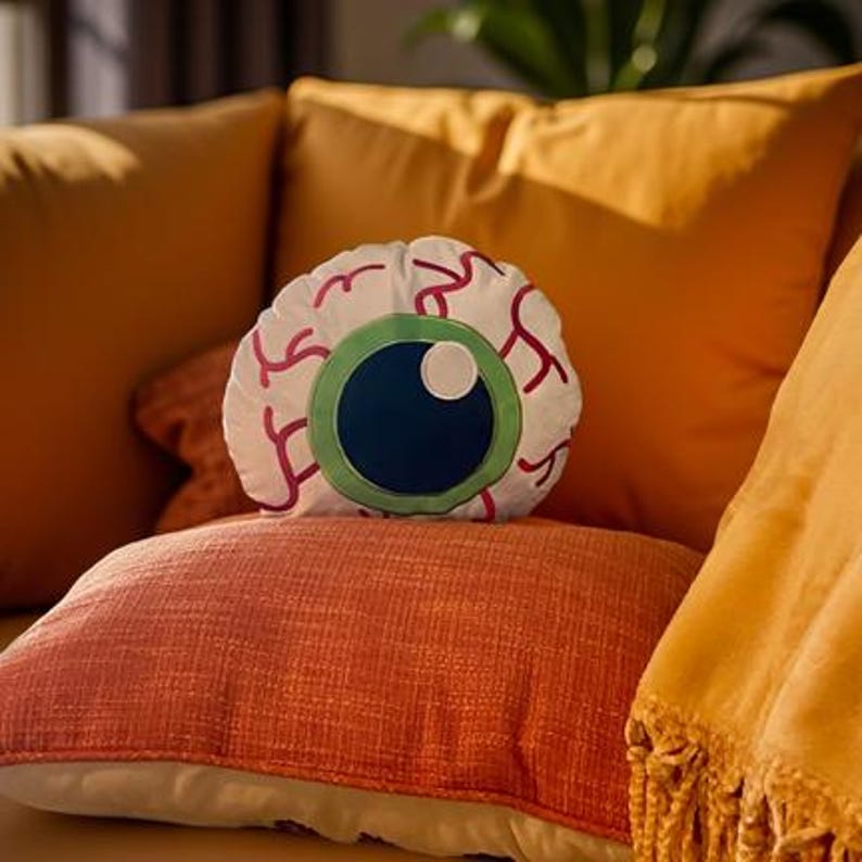 May include: A white plush pillow shaped like a giant eyeball with a green iris and a black pupil. The pillow is sitting on a couch with orange and yellow cushions.