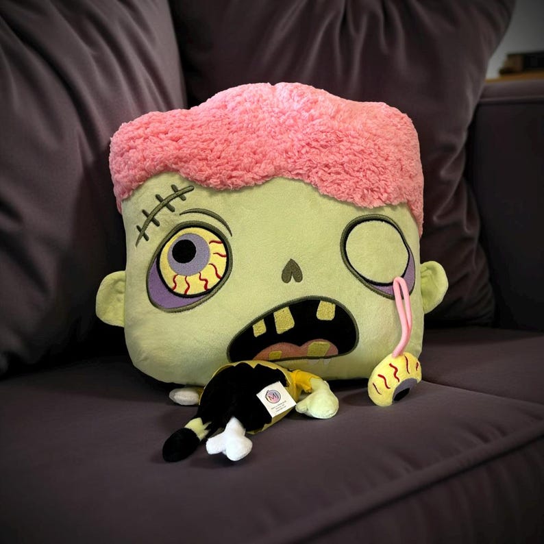 May include: A green and pink plush zombie pillow with a stitched-up mouth, one eye missing and a dangling eyeball. The zombie is holding a small plush black and yellow creature with a bone.