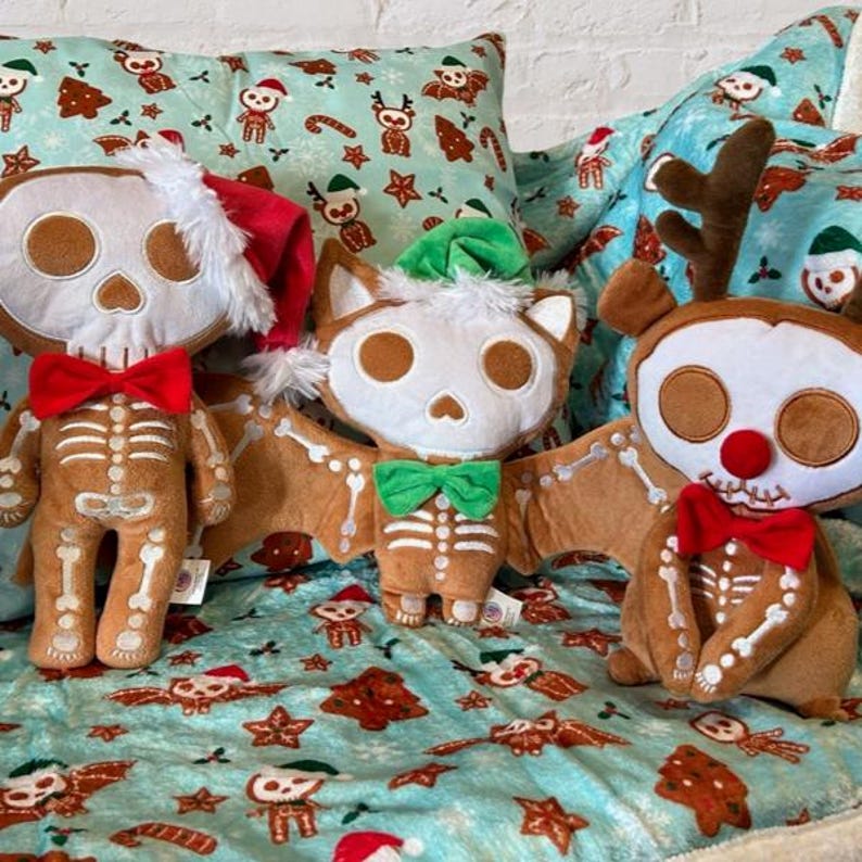 Op de afbeelding: Drie gemberkoek skelet-knuffels met verschillende feestthema's. Een skelet met een kerstmanmuts, een skelet-vleermuis met een groene hoed en een rendierskelet met een rode muts.