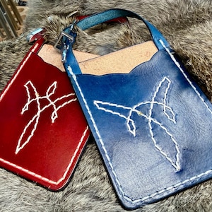 May include: Two leather phone cases, one red and one blue, with white stitched designs. Each case has a strap and a unique white stitched design. The cases are displayed on a textured, fur-like surface.