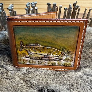 May include: A brown leather wallet with a hand-painted image of a fish in a natural setting. The wallet has a bi-fold design with white stitching around the edges. The fish is yellow and brown, set against a green and brown background.