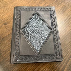 May include: A dark gray leather journal with a diamond-shaped, textured, light gray panel in the center. The journal has a decorative border and is closed. The cover is made of leather.