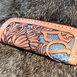 May include: A handcrafted leather wallet with intricate floral and scrollwork carving. The wallet features a light brown exterior with a blue-green interior and decorative stitching. The design includes the number "50" in a stylized font.