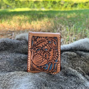 May include: A brown leather wallet with a detailed floral and bird design. The wallet has a light brown border and a blue background within the carved design. The wallet is sitting on a gray fur surface.