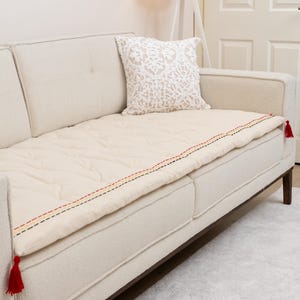 Puede incluir: Funda de sofá acolchada de color crema con un borde cosido decorativo en rojo, negro y amarillo. La funda tiene borlas rojas en cada extremo. Un cojín estampado descansa sobre el sofá. El sofá tiene patas de madera oscura.