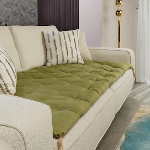 May include: A cream sofa with an olive green, tufted cushion and patterned pillows. A gold floor lamp with spherical details is visible. A tassel hangs from the sofa arm.