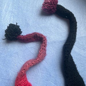May include: Two handmade knit hat tails with pom-poms. One tail is black with a dark pink pom-pom, and the other is a gradient of pink and red with a black pom-pom. The tails are on a light blue background.