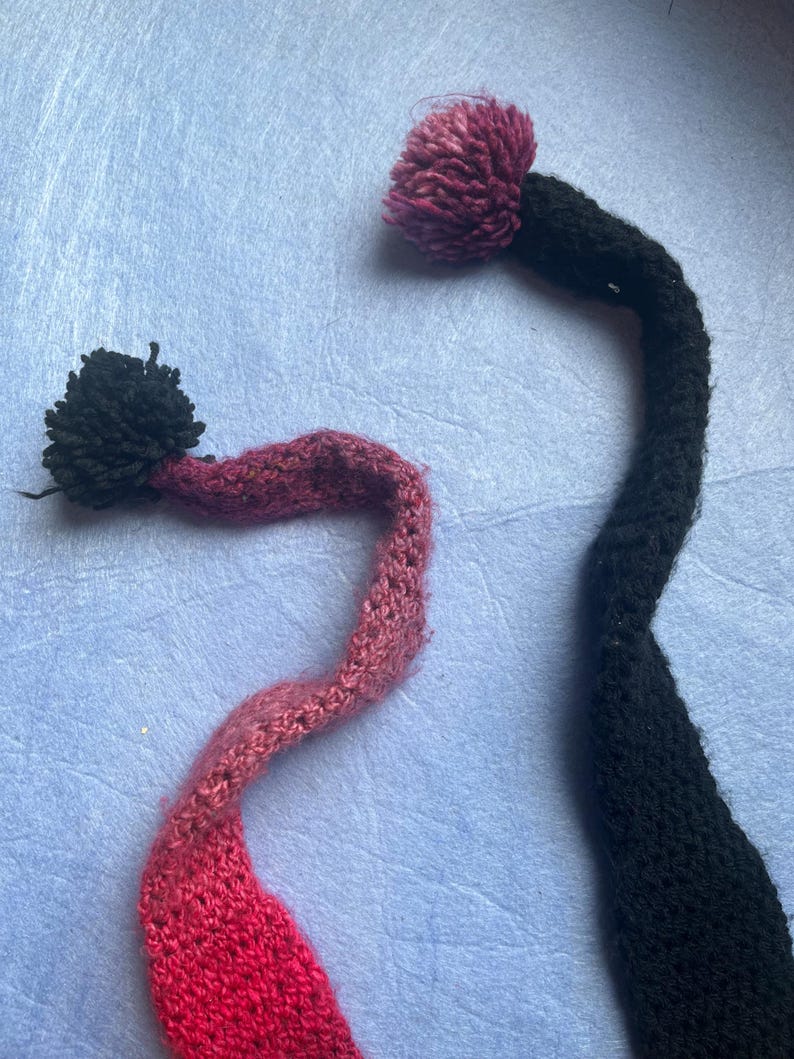 May include: Two handmade knit hat tails with pom-poms. One tail is black with a dark pink pom-pom, and the other is a gradient of pink and red with a black pom-pom. The tails are on a light blue background.