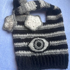 May include: A handmade crochet tote bag in black, gray, and white. The bag features horizontal stripes and a central eye design. The handle and decorative elements are also crocheted in coordinating colors. The bag is a unique, handcrafted accessory.