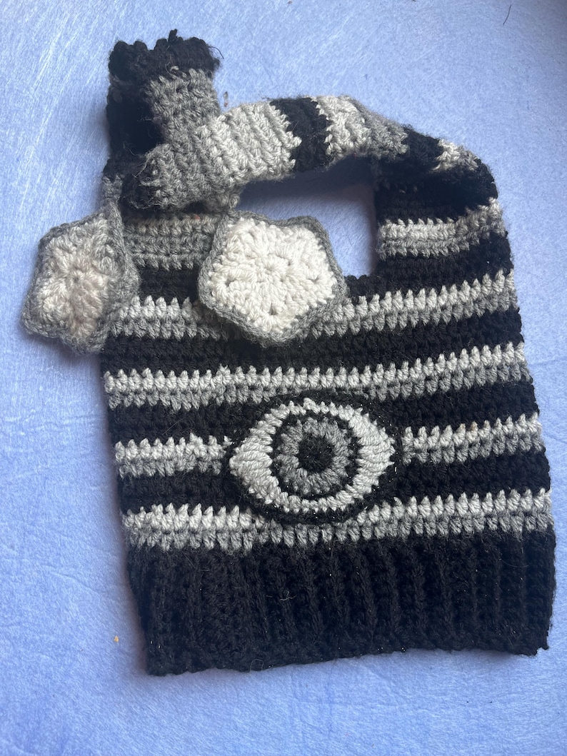 May include: A handmade crochet tote bag in black, gray, and white. The bag features horizontal stripes and a central eye design. The handle and decorative elements are also crocheted in coordinating colors. The bag is a unique, handcrafted accessory.
