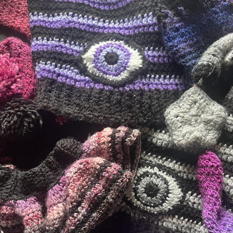 May include: A collection of handmade crochet items in various colors. The items include a black hat with a purple and white eye design, mittens, and other accessories. The crochet work features a variety of textures and patterns.